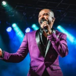 Alexandre Pires canta ao microfone no palco, de paletó roxo com lapela preta, sob luzes azuis e fumaça