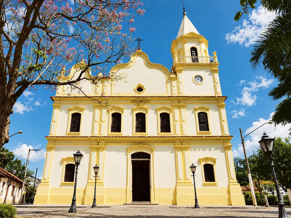 Fachada frontal da Igreja Matriz de Sant'Ana, amarela e branca, com torre de relógio e árvore florida à frente