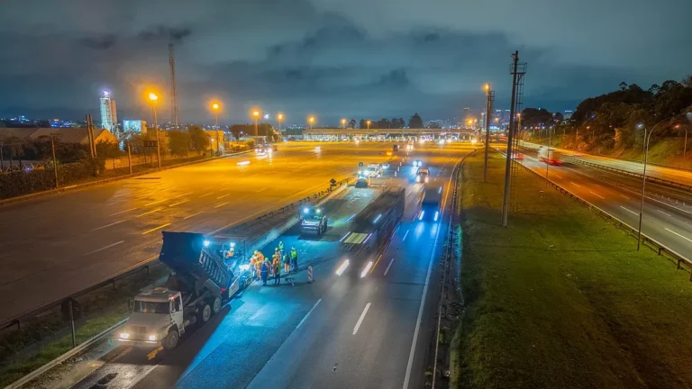 Equipe faz recapeamento noturno em pista de rodovia, com caminhão basculante e praça de pedágio ao fundo