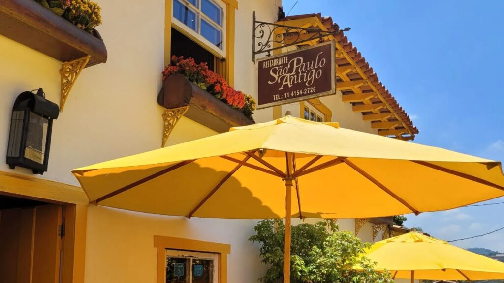 Fachada amarela do casarão do São Paulo Antigo Restaurante, com placa, flores e mesas sob ombrelones, em Santana de Parnaíba