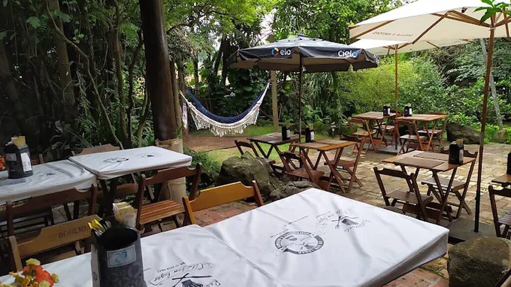 Área externa do Restaurante Um Bom Lugar, com mesas de madeira, ombrelones e uma rede entre as árvores, em Santana de Parnaíba